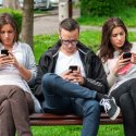 Three young men and women sitting on park bench looking at their phones